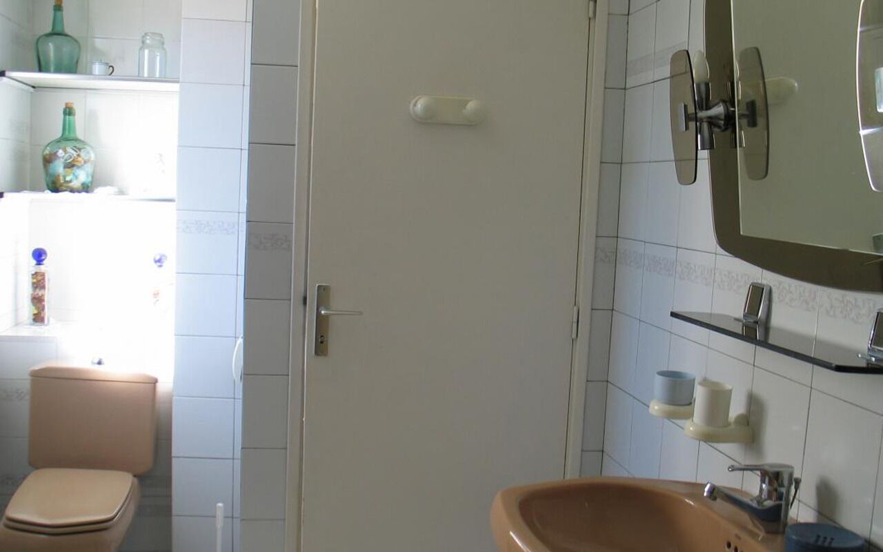 A bathroom with a toilet, sink, and mirror. There are shelves with toiletries and a window with natural light.