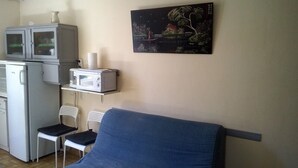Uma pequena cozinha compacta com micro-ondas, geladeira e armário. Há uma cadeira azul e uma cadeira branca. Um quadro está na parede.