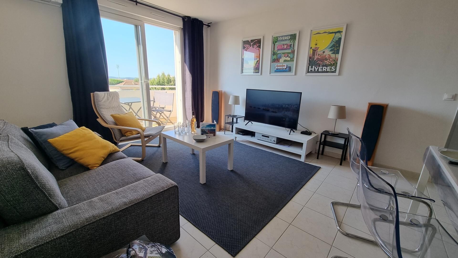 Een moderne woonkamer met een grijze bank, gele kussens, een salontafel, een televisie en een balkon met buitenmeubilair.