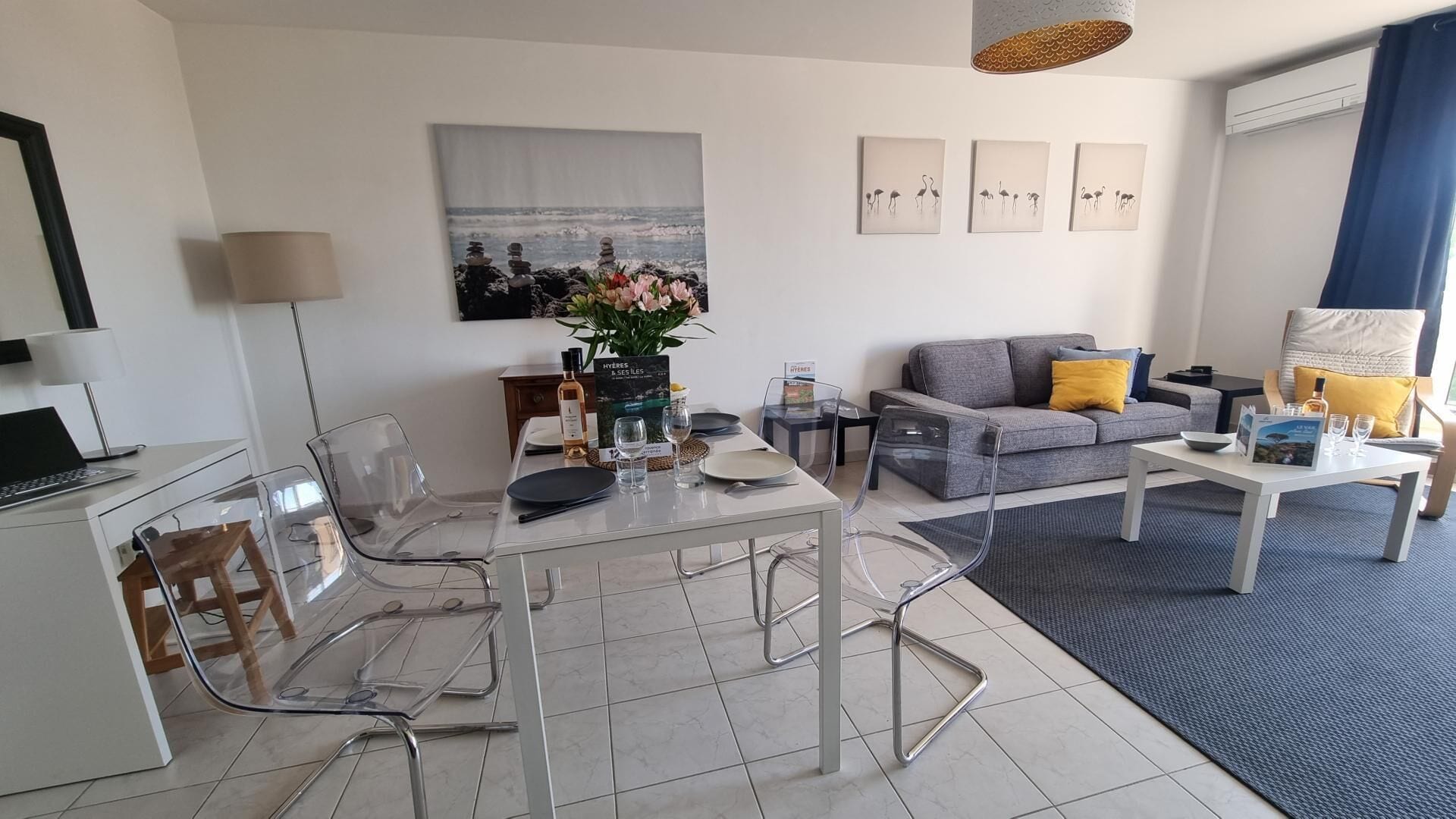 A modern living room with a dining area, featuring a grey sofa, clear chairs, and a dining table set for two.