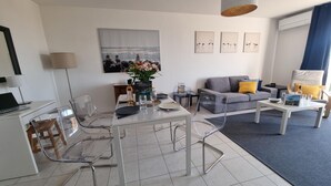 Una sala de estar moderna con comedor, que incluye un sofá gris, sillas transparentes y una mesa de comedor para dos personas.