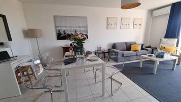 A modern living room with a dining area, featuring a grey sofa, clear chairs, and a dining table set for two.