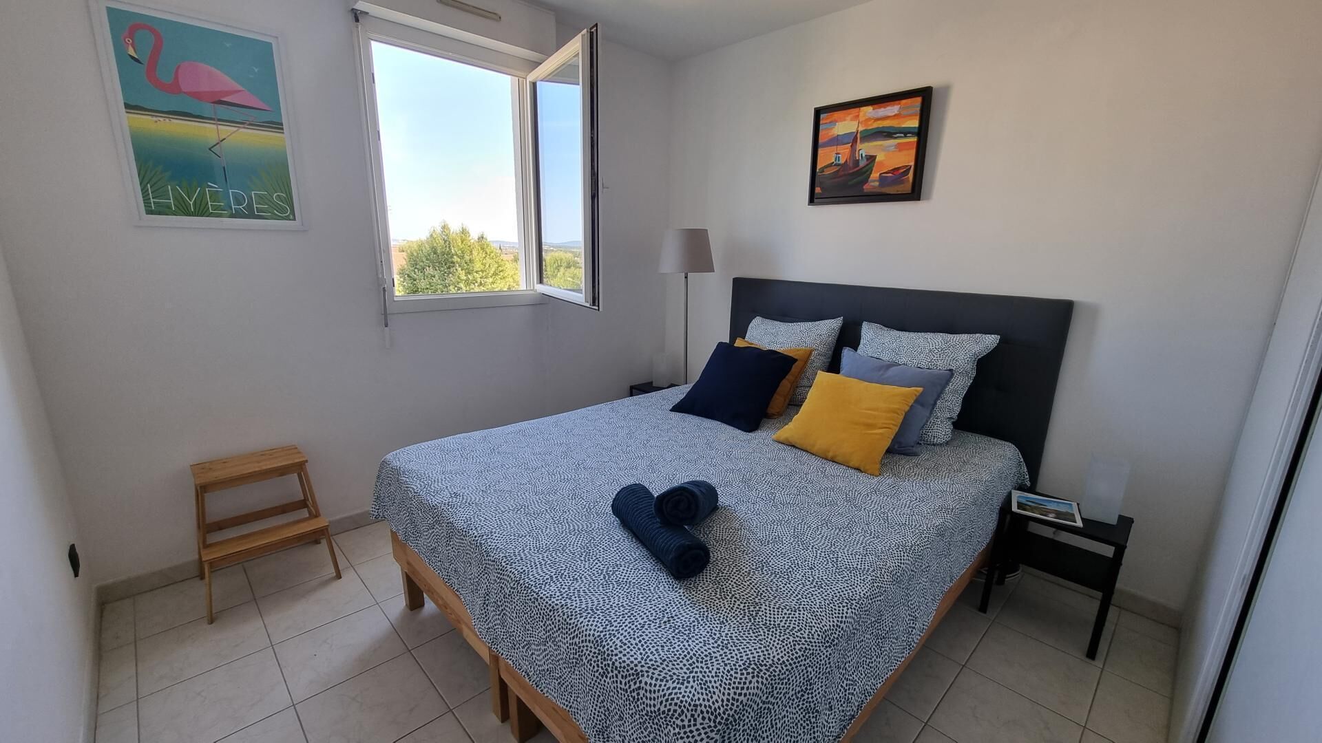 A bedroom with a bed, pillows, a nightstand, a lamp, a small wooden stool, and two paintings on the wall.