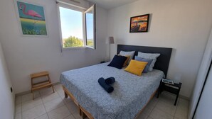 Un dormitorio con cama, almohadas, una mesita de noche, una lámpara, un taburete de madera y dos cuadros en la pared.