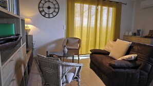 Un salon avec un canapé marron, un fauteuil en osier, une petite table, une horloge au mur et un rideau jaune.