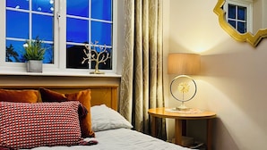 A hotel room with a bed, a nightstand with a lamp, a window with curtains, and a mirror on the wall. - Lake house - 3 bedroom woking (Woking)