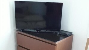 Une pièce avec une commode marron adossée à un mur blanc, un téléviseur à écran plat posé sur le dessus, et une chaise en bois.