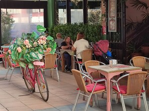 Ett utomhuskafé med flätade stolar och bord, en cykel med en blomkorg och en grön häck i bakgrunden.