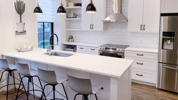 A modern kitchen with white cabinetry, a central island with bar stools, stainless steel appliances, pendant lighting, and a view to the outdoors. - Downtown Heritage Condo (Charlottetown)
