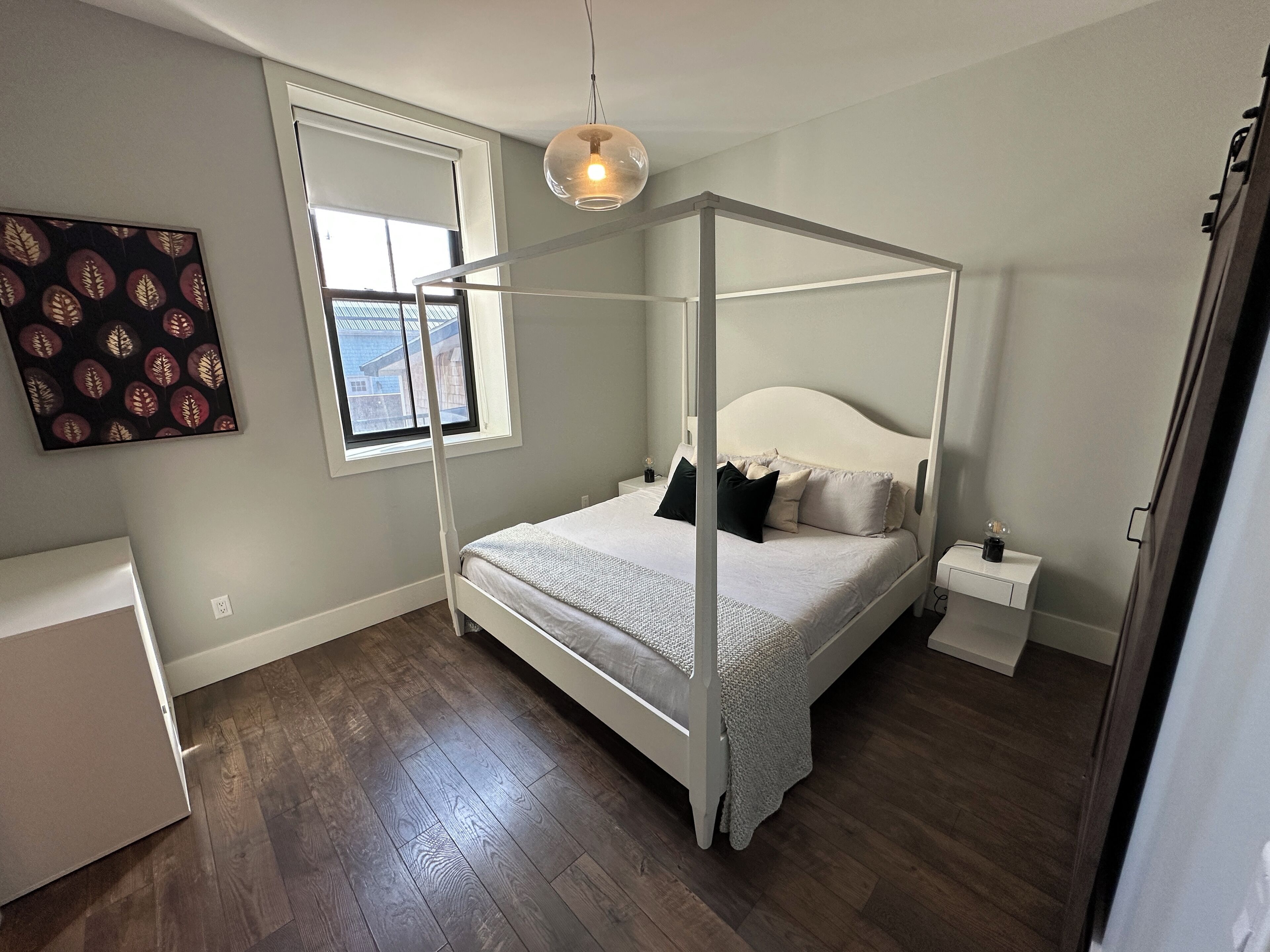 A bedroom with a four-poster bed, a bedside table, a nightstand, a window, and a wall-mounted artwork.
