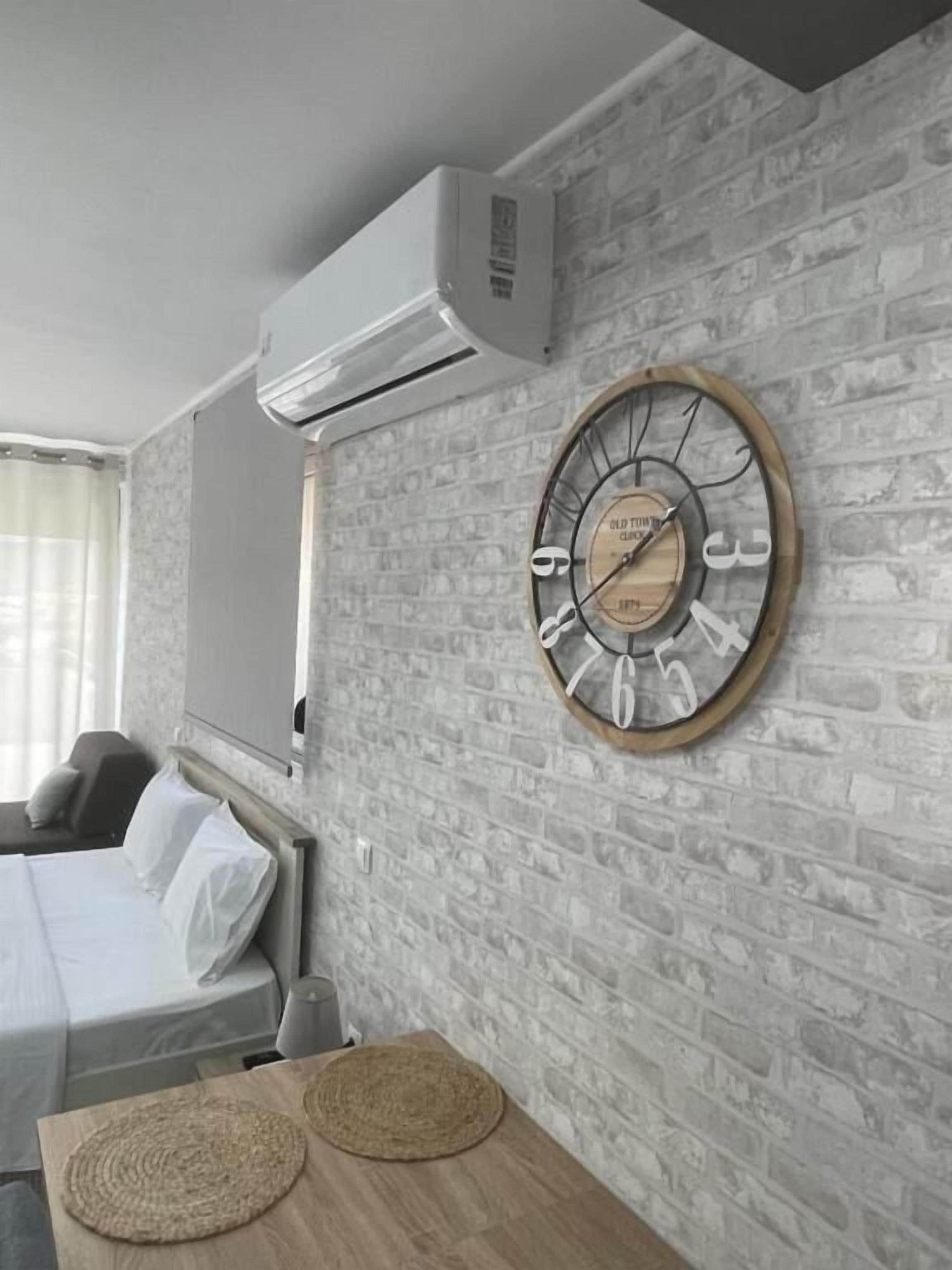 Una habitación de hotel con pared de ladrillo, un reloj de madera redondo, una cama con ropa blanca y una pequeña mesa con dos individuales tejidos.
