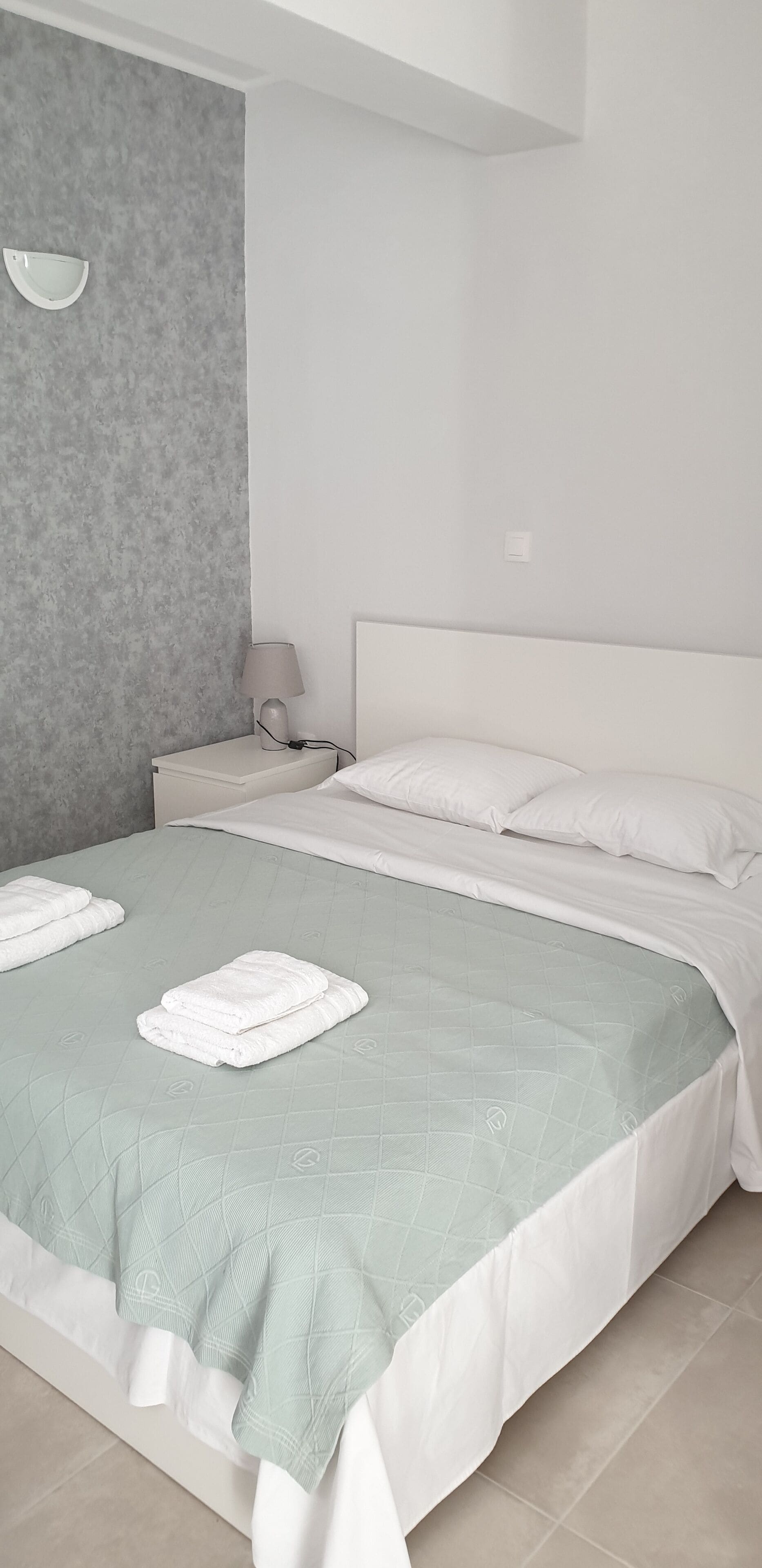Uma cama bem arrumada com lençóis brancos e um edredom verde-claro, uma luminária de cabeceira e uma mesa de cabeceira com abajur.