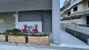 A modern building with a closed shutter, red chairs, and wooden planters with greenery. - AVR DELUXE SUITES 6 AIRPORT FREE SHUTTLE (Markopoulo Mesogaias)