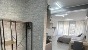A modern living room with a built-in wardrobe, a dining area with a wooden table and chairs, and a large window with sheer curtains. - AVR DELUXE SUITES 6 AIRPORT FREE SHUTTLE (Markopoulo Mesogaias)