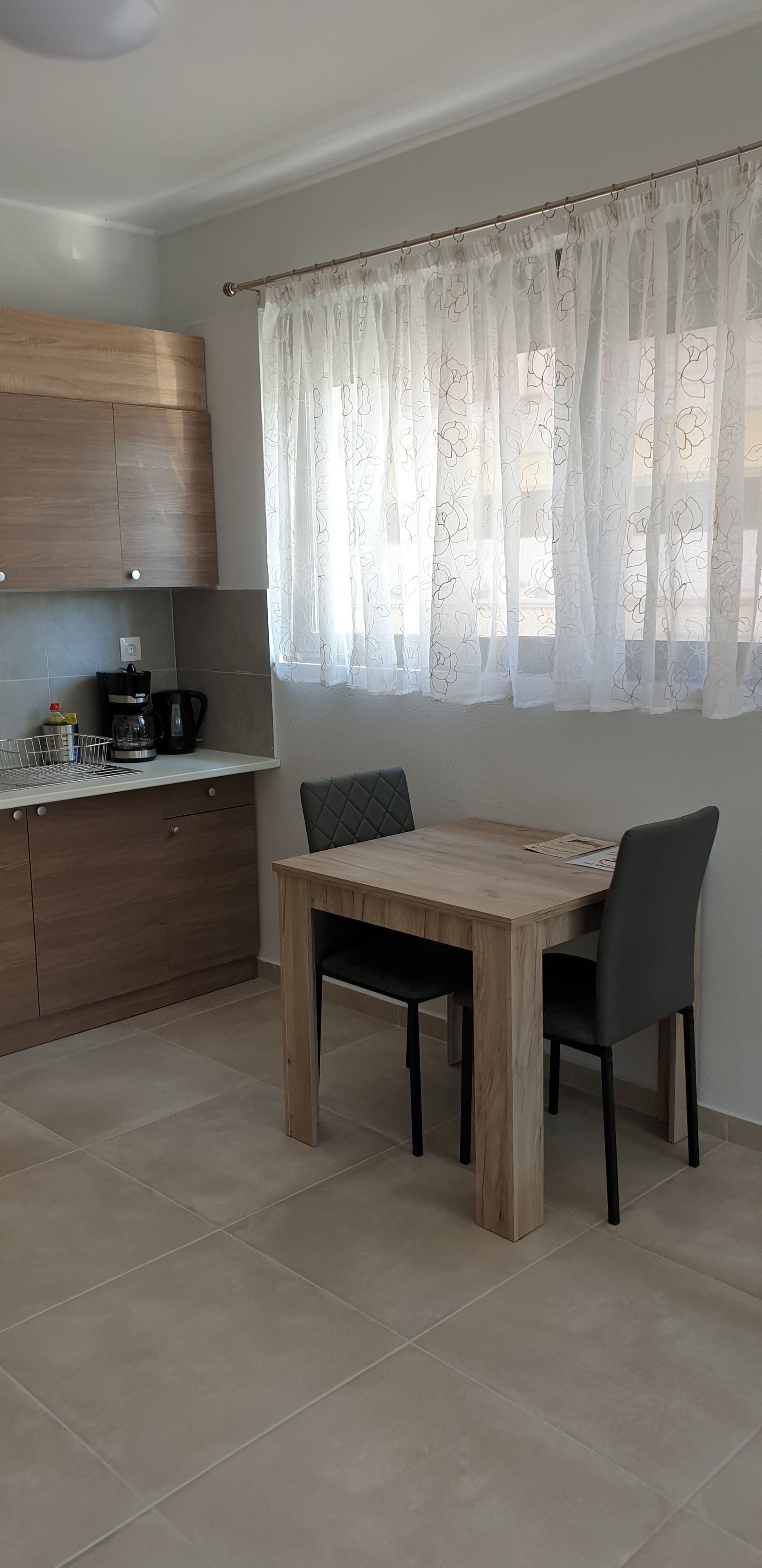 A modern kitchen with wooden cabinets, a dining table with chairs, and a window with sheer curtains.