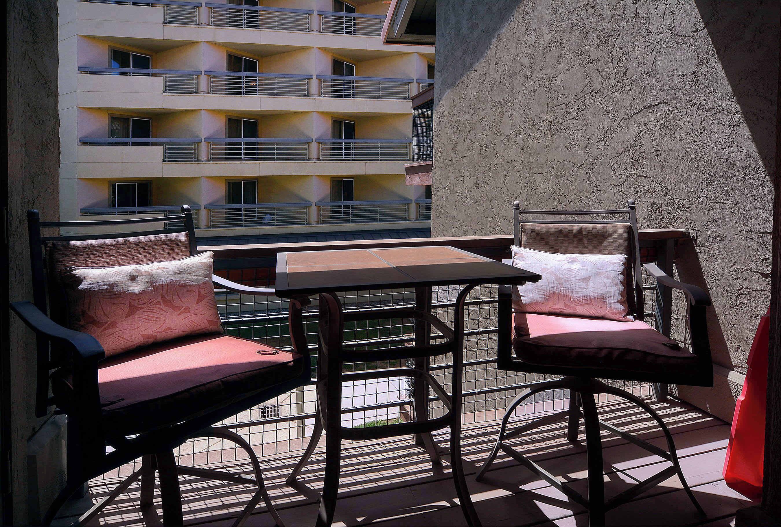 An outdoor balcony with a table and two chairs, a textured wall, and a building with balconies in the background.