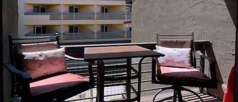 An outdoor balcony with a table and two chairs, a textured wall, and a building with balconies in the background.