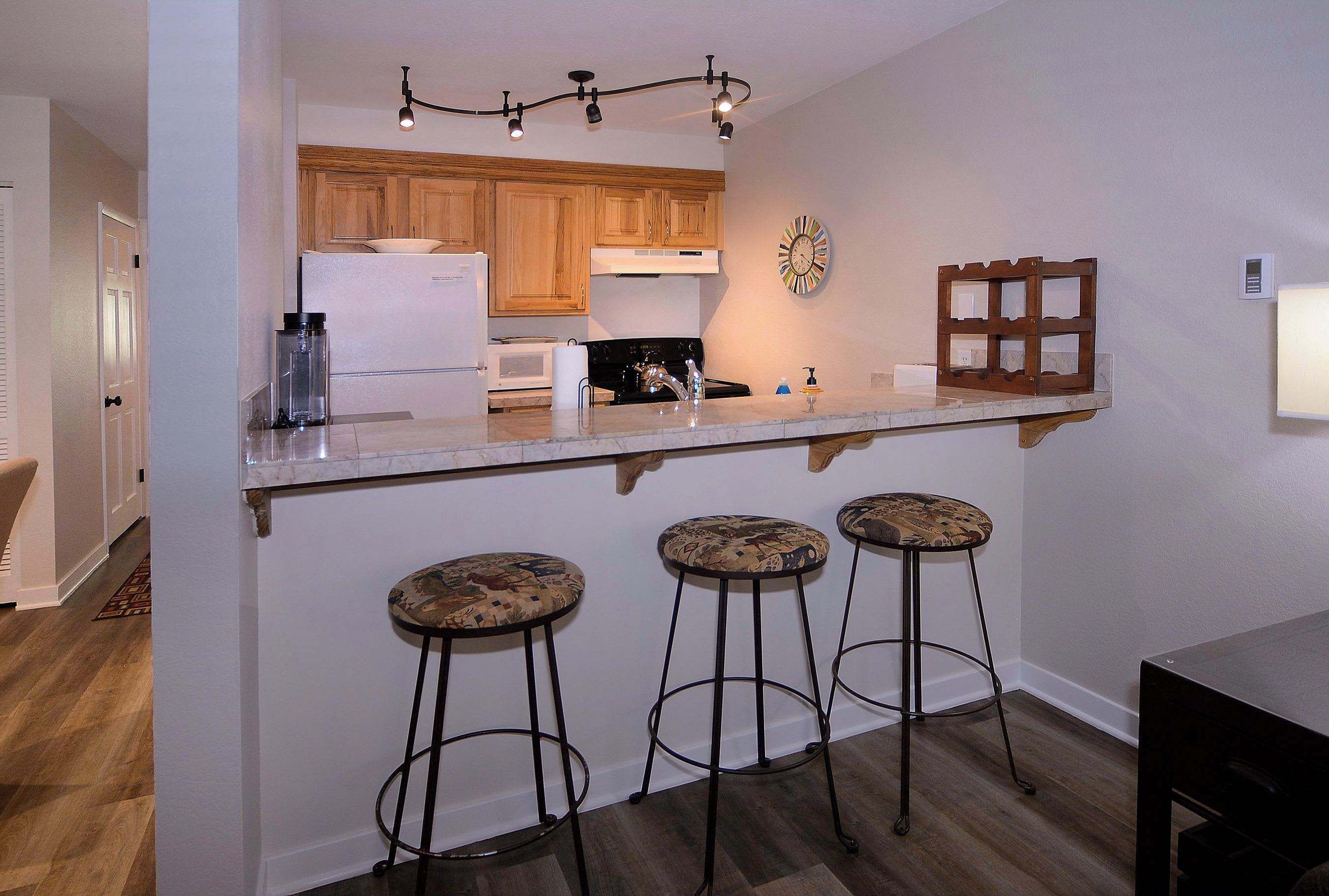 A kitchen with wooden cabinets, a white refrigerator, a microwave, and a sink. There are three stools with patterned seats.