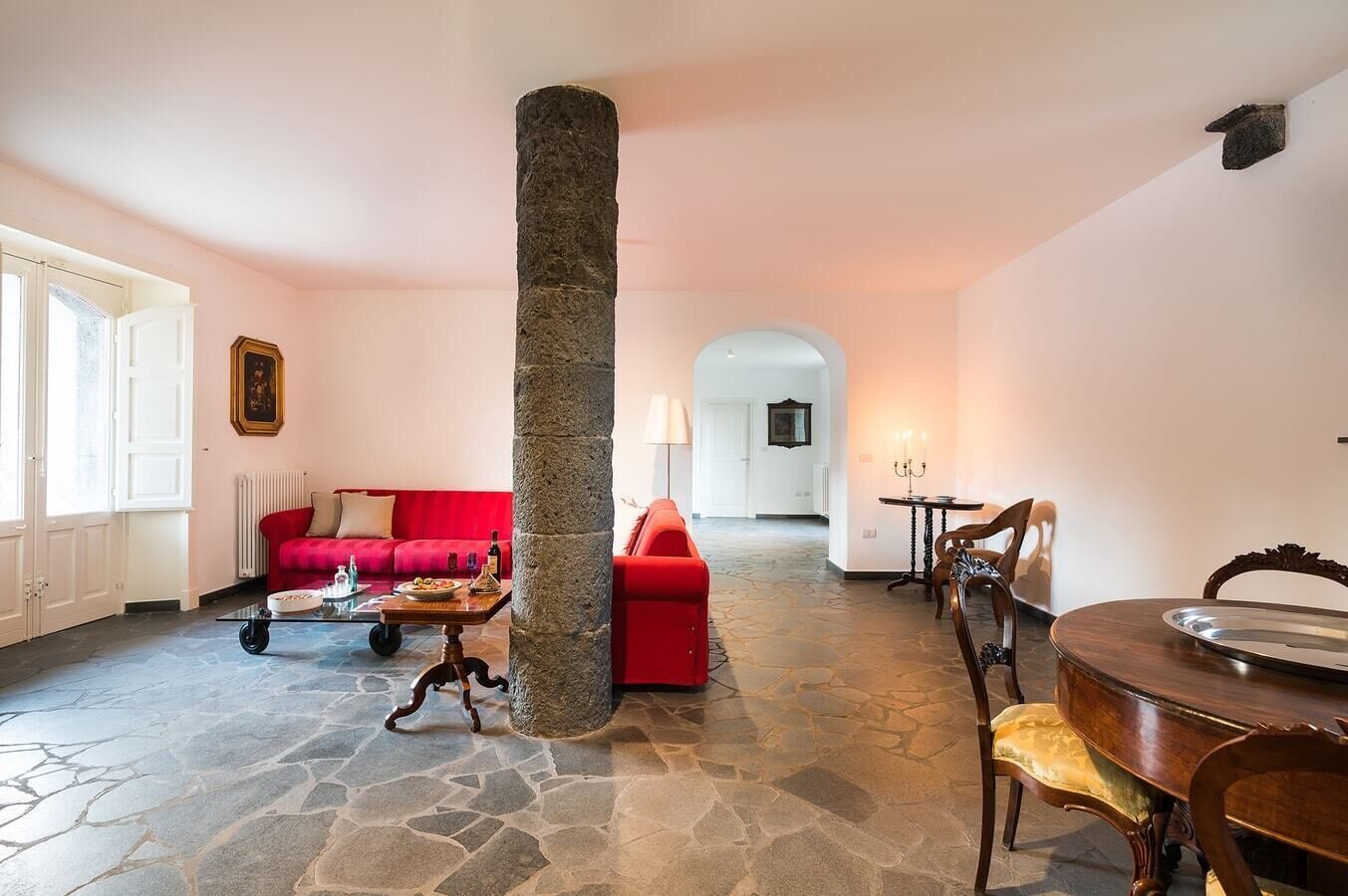 Uma sala de estar espaçosa com piso de pedra, um sofá vermelho, uma mesa de centro de madeira e uma mesa de jantar com cadeiras.