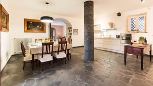 A dining area with a stone floor, a central pillar, and a kitchen in the background. - Villa Coco - 10 Guests, Stunning Mt Etna Views (Castiglione di Sicilia)