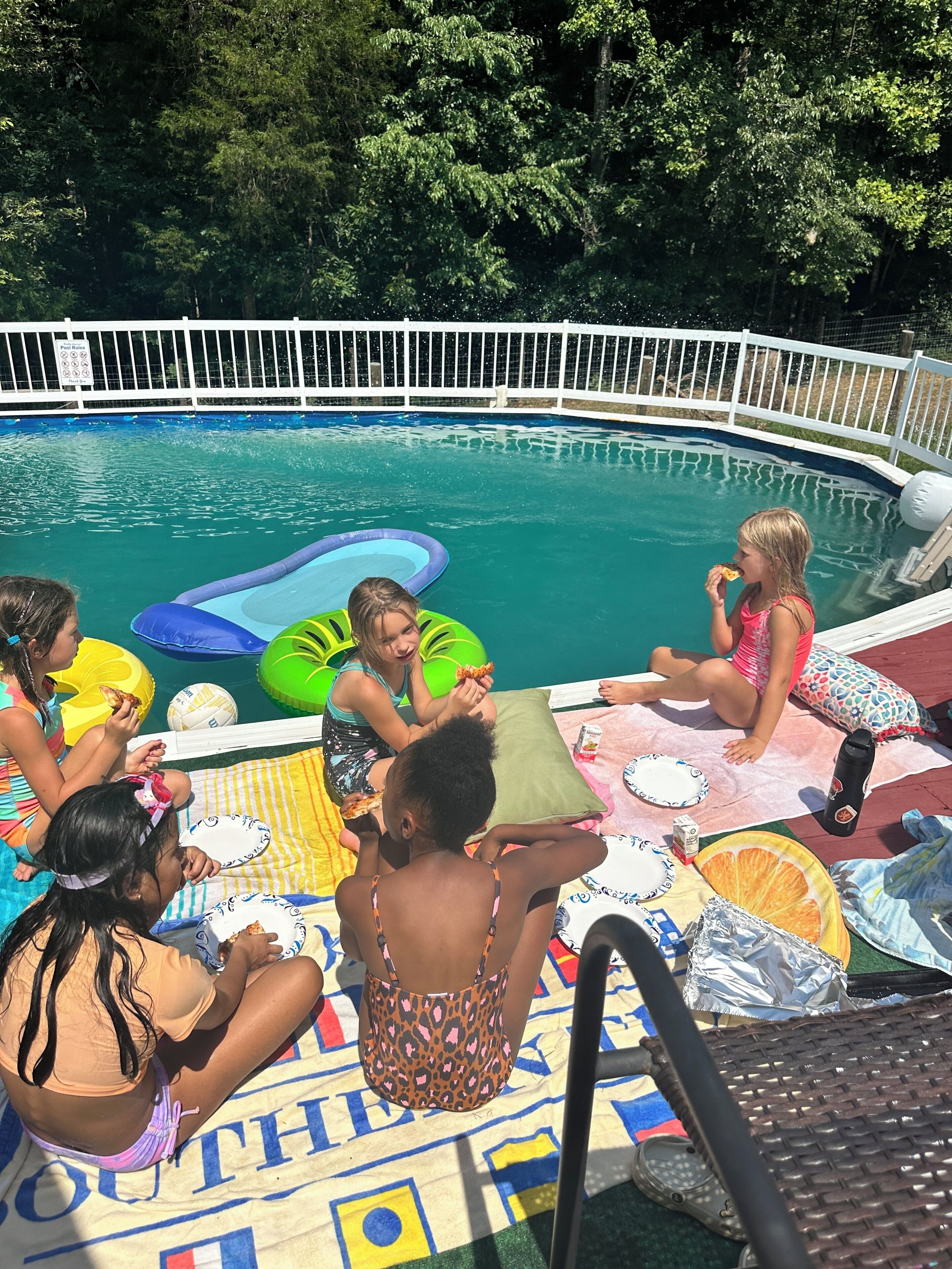 Une piscine avec des jeux gonflables, un espace de détente au bord de la piscine avec des serviettes et un parasol, ainsi qu’une clôture autour de la piscine.
