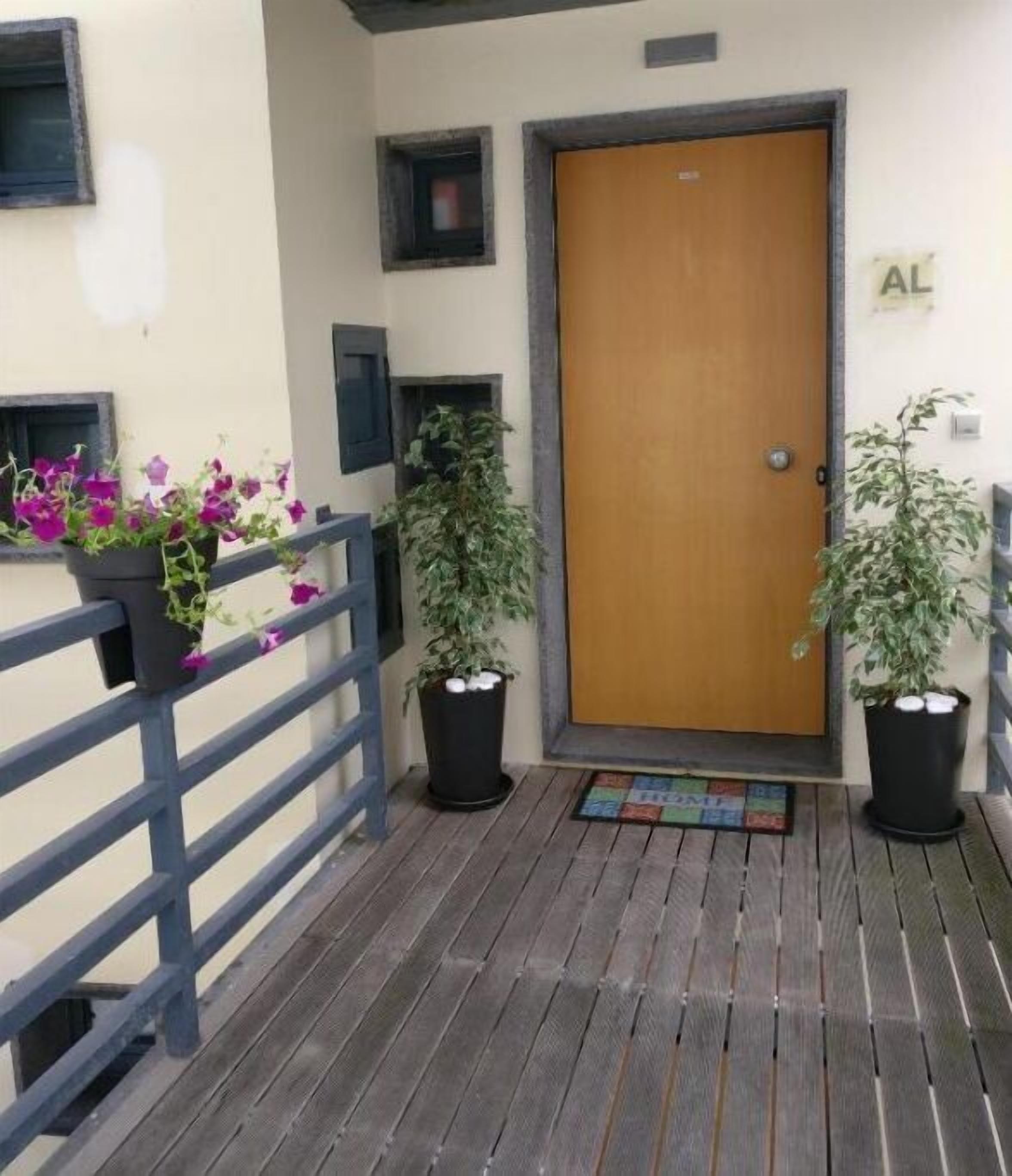 Une terrasse en bois avec une rambarde en métal, une porte en bois et deux plantes en pot ornées de fleurs.
