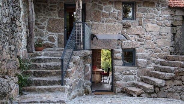 Edifici de pedra amb una escala de pedra que condueix a una porta, una petita zona d'estar a l'exterior amb una taula i cadires, i una finestra amb vista a vegetació.