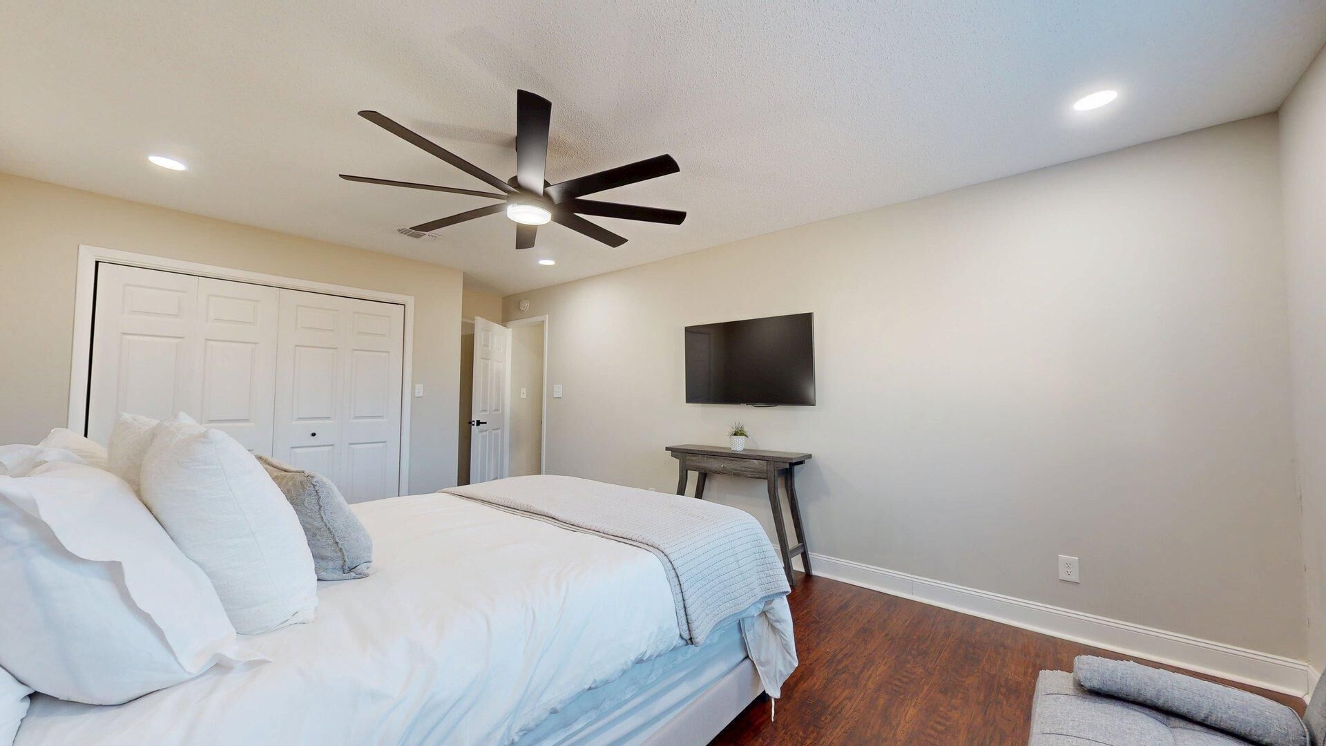A bedroom with a bed, a ceiling fan, a TV on a stand, and a door.
