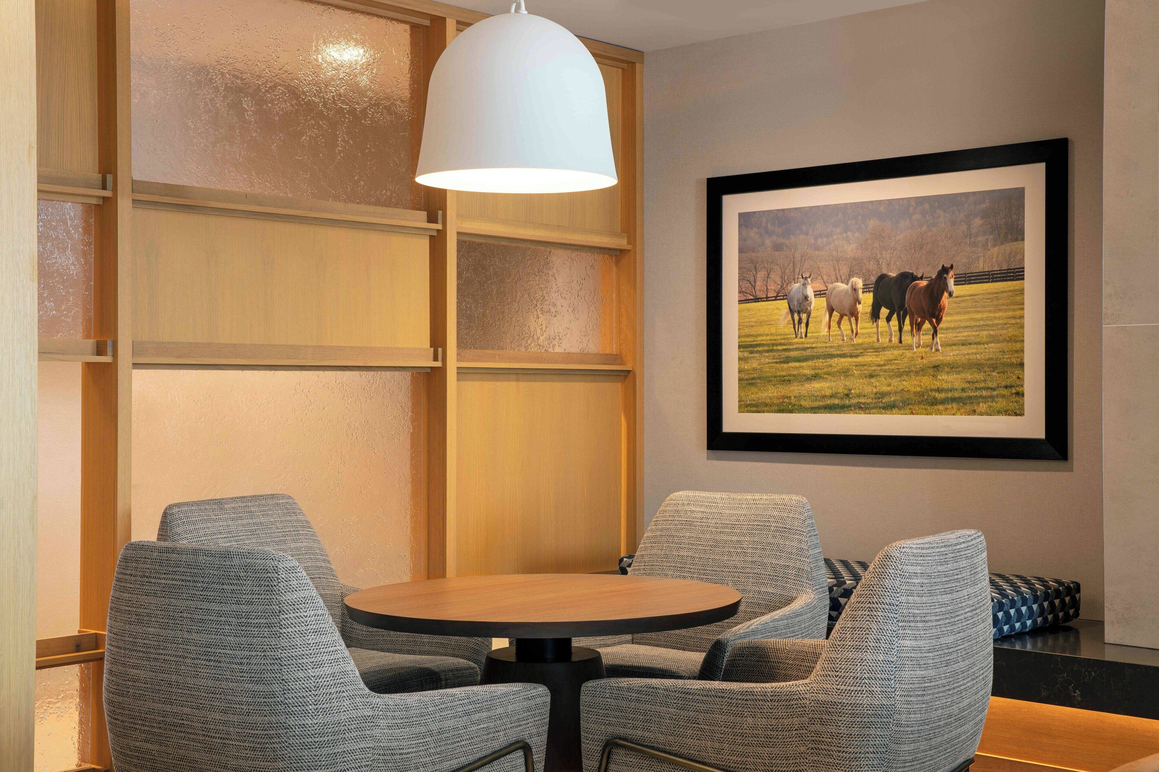 Una habitación con una mesa redonda, cuatro sillas grises tapizadas, una lámpara colgante blanca y un cuadro con caballos en la pared.