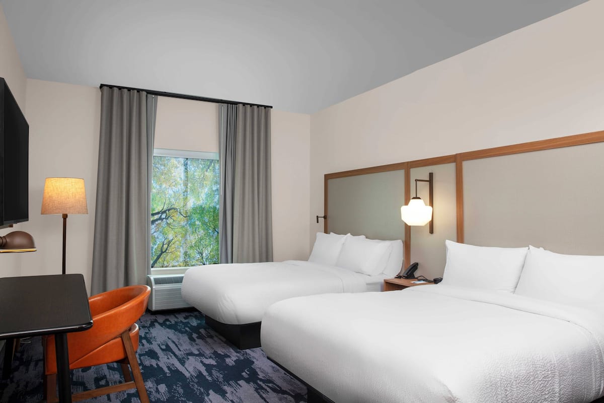 Une chambre d’hôtel avec deux lits, un bureau, une chaise orange, une lampe et une fenêtre avec des rideaux.
