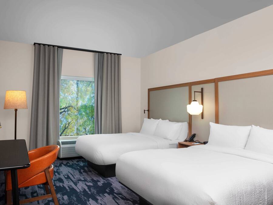 A hotel room with two beds, a desk, an orange chair, a lamp, and a window with curtains.