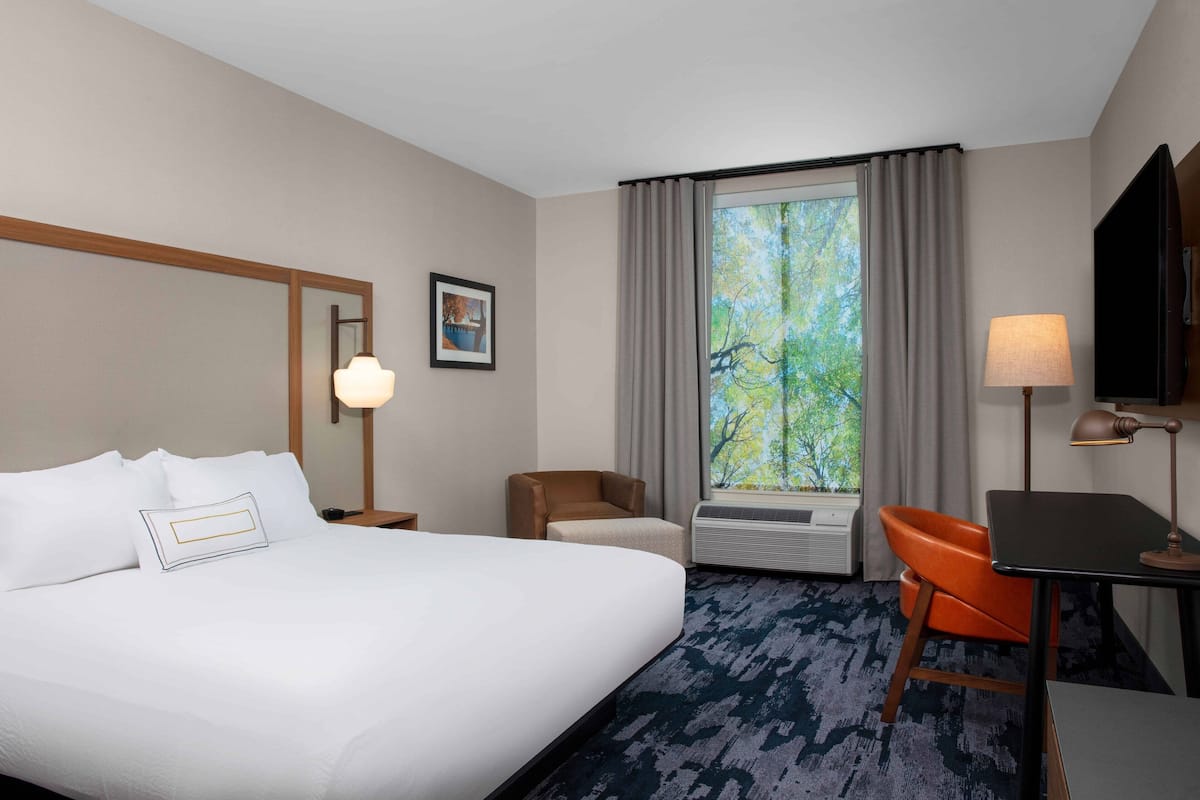 Une chambre d’hôtel avec un grand lit, un bureau, une chaise, une fenêtre avec des rideaux, un téléviseur fixé au mur et un tableau encadré.