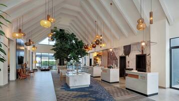 A spacious lobby with a high ceiling, hanging wicker pendant lights, a central indoor plant, a reception desk, and a large window providing natural light.