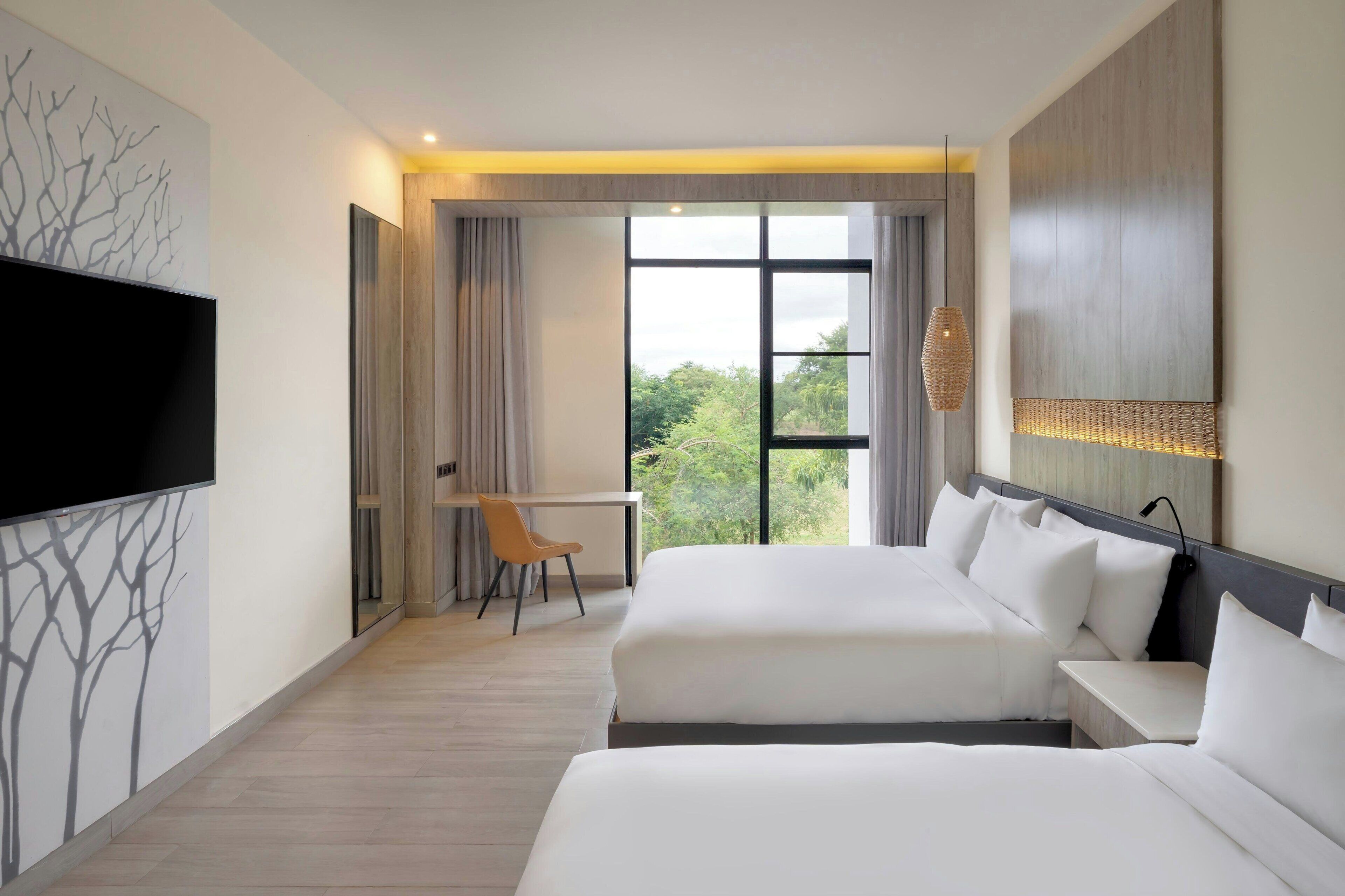 A hotel room with two beds, a desk, a chair, a TV, and a large window with a view.