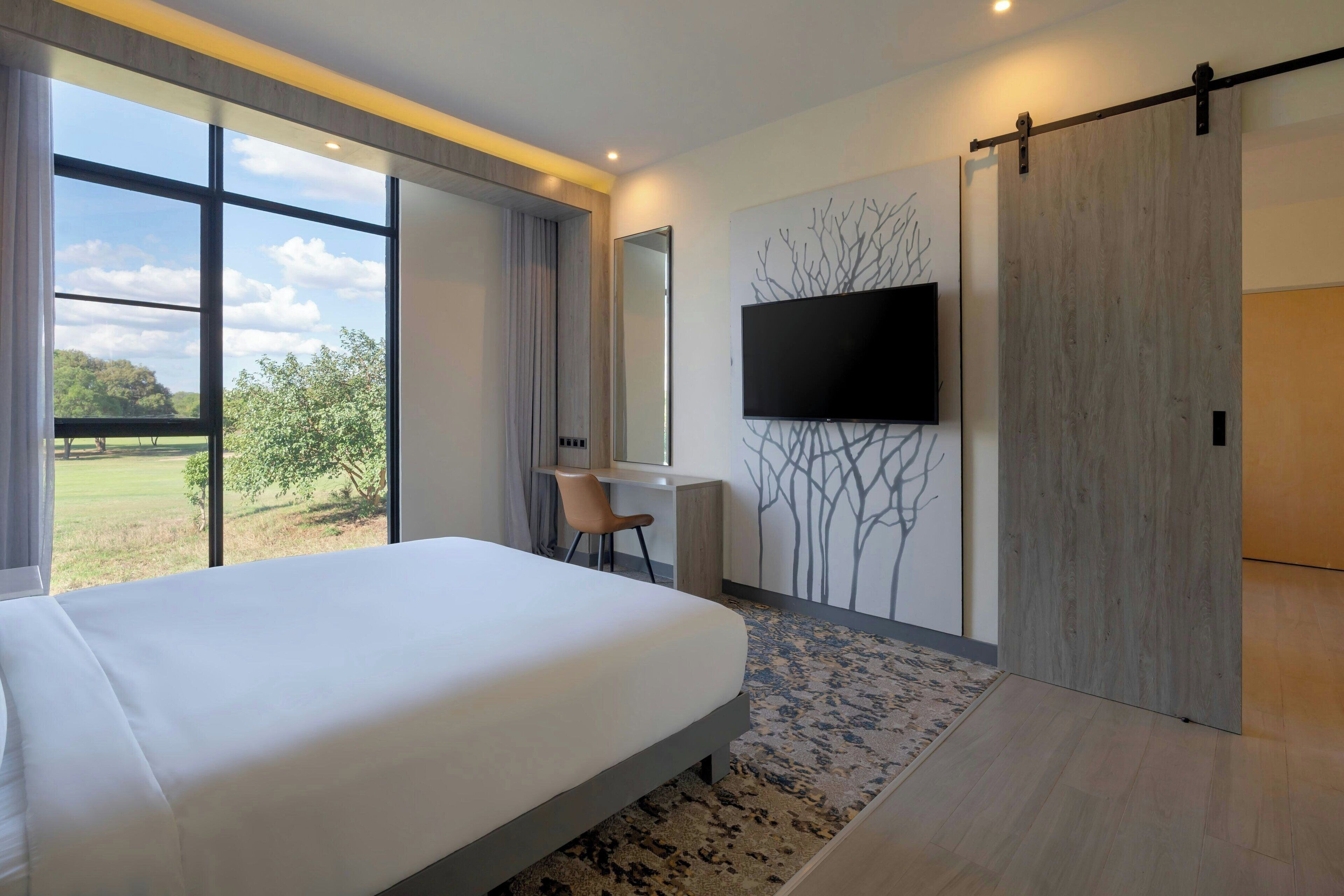 Una camera d'albergo moderna con un letto grande, una scrivania con una sedia, una TV a parete e una porta scorrevole.