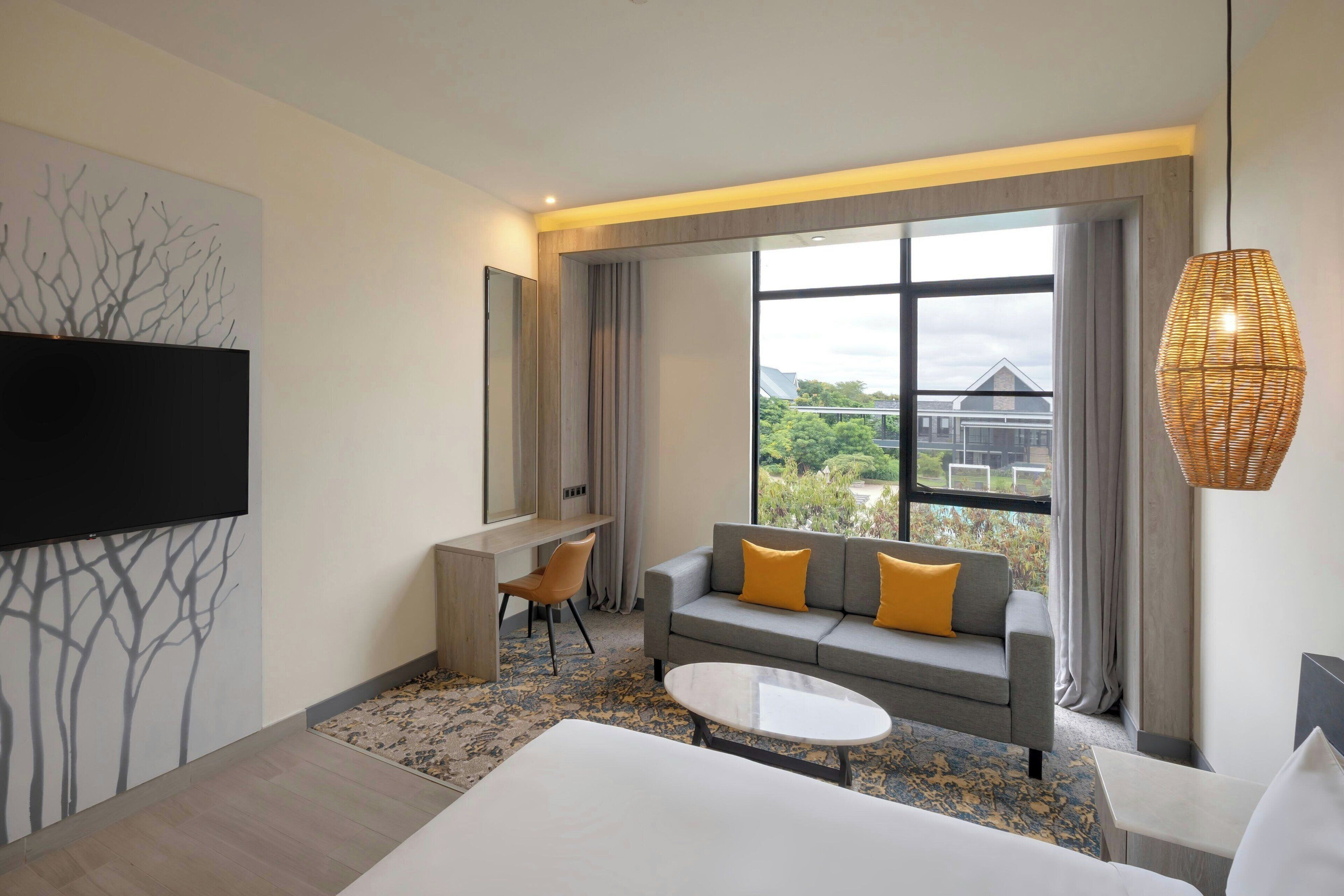 Una habitación de hotel moderna con un ventanal grande, un sofá con cojines amarillos, un televisor de pantalla plana montado en la pared y un escritorio pequeño.