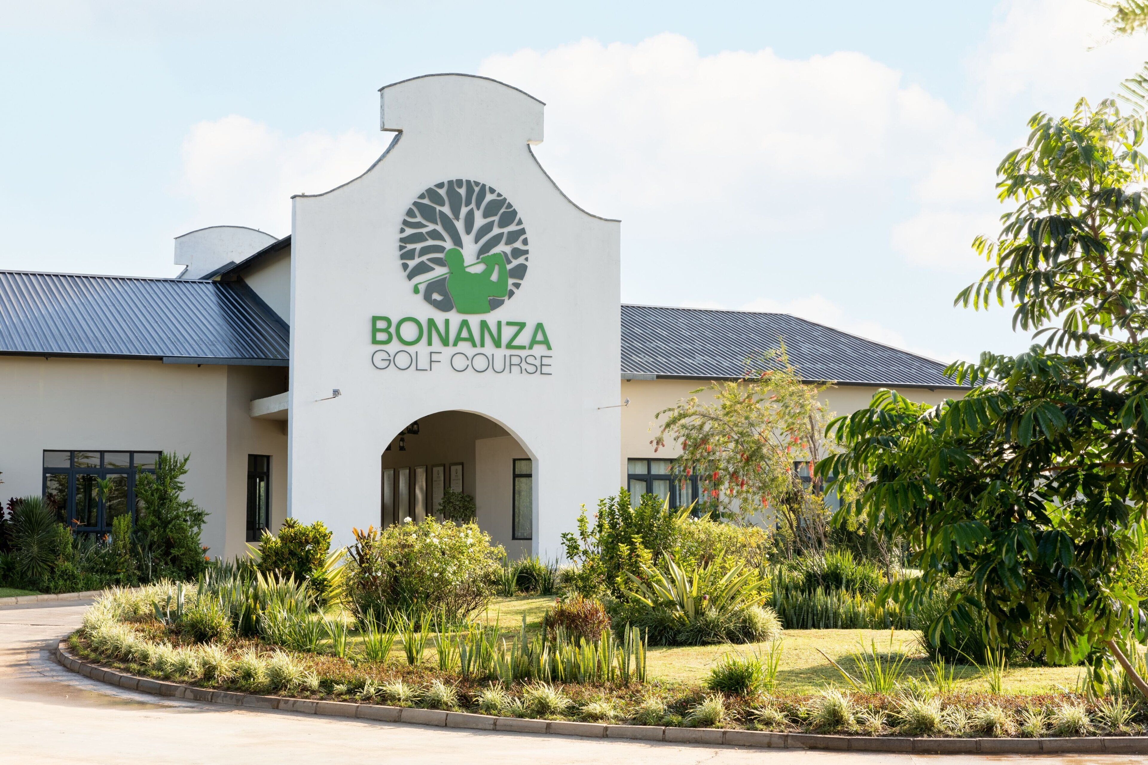 A golf course entrance with a sign, a building with a sloped roof, and landscaped gardens.