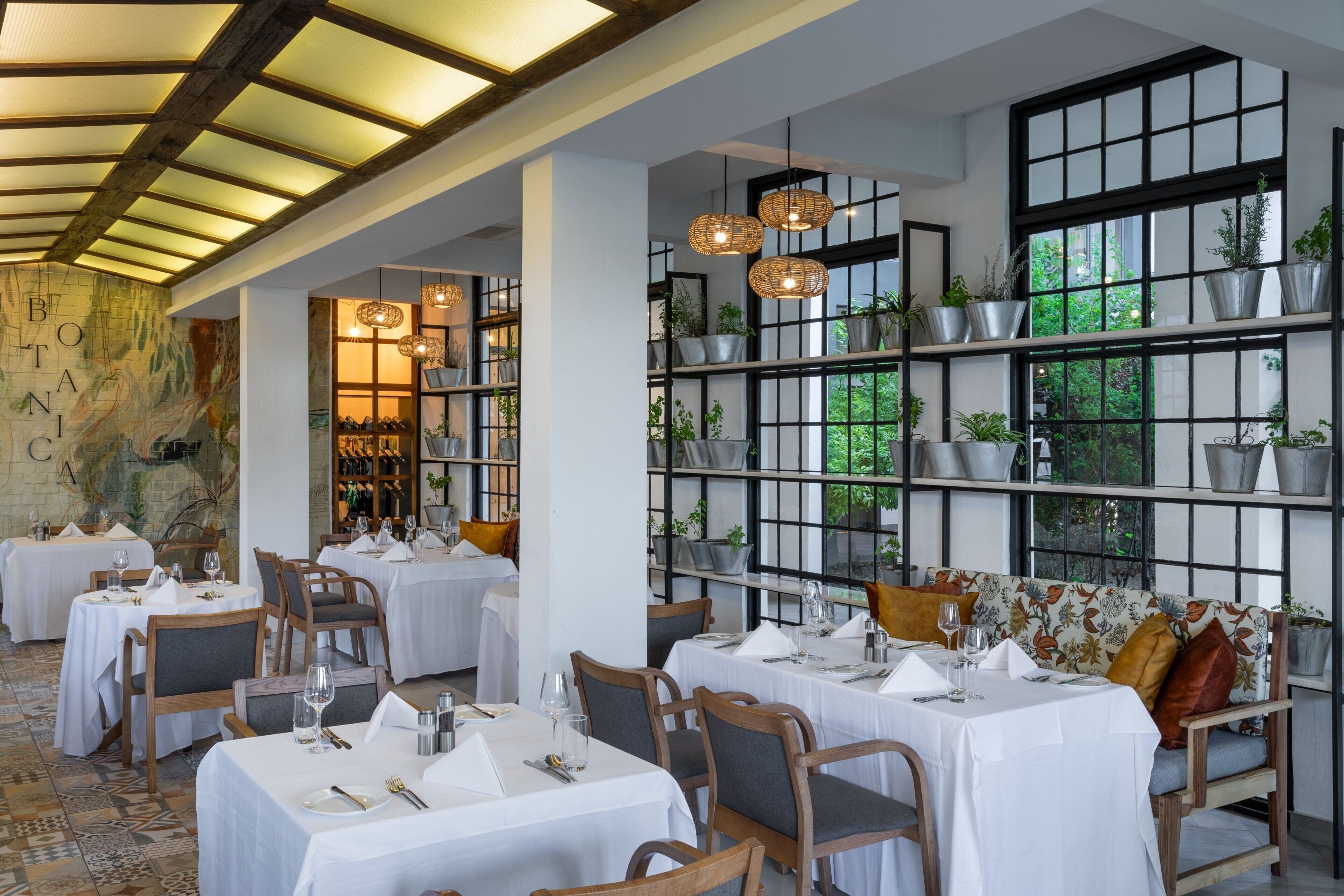 A restaurant with tables set for dining, large windows with plants, and a ceiling with exposed beams.