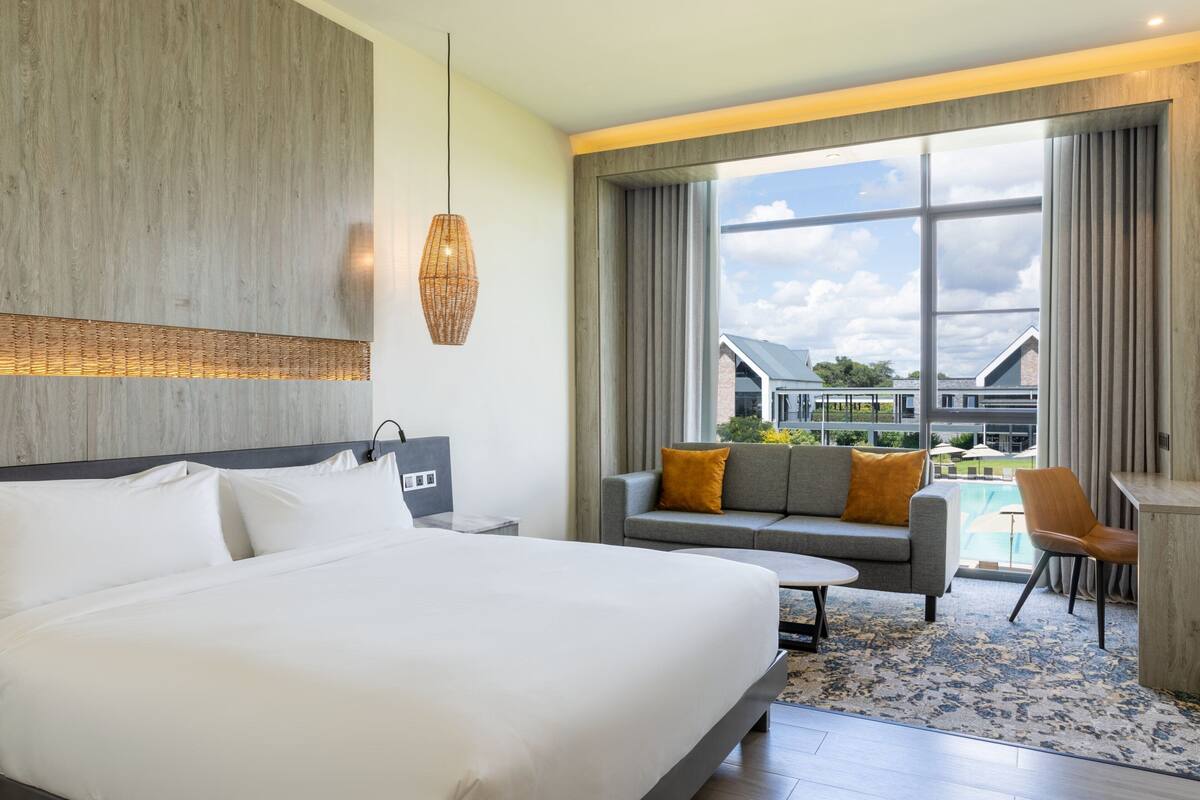 Ein modernes Hotelzimmer mit einem großen Bett, einer Couch, einem Schreibtisch und Blick auf einen Swimmingpool und Gebäude.