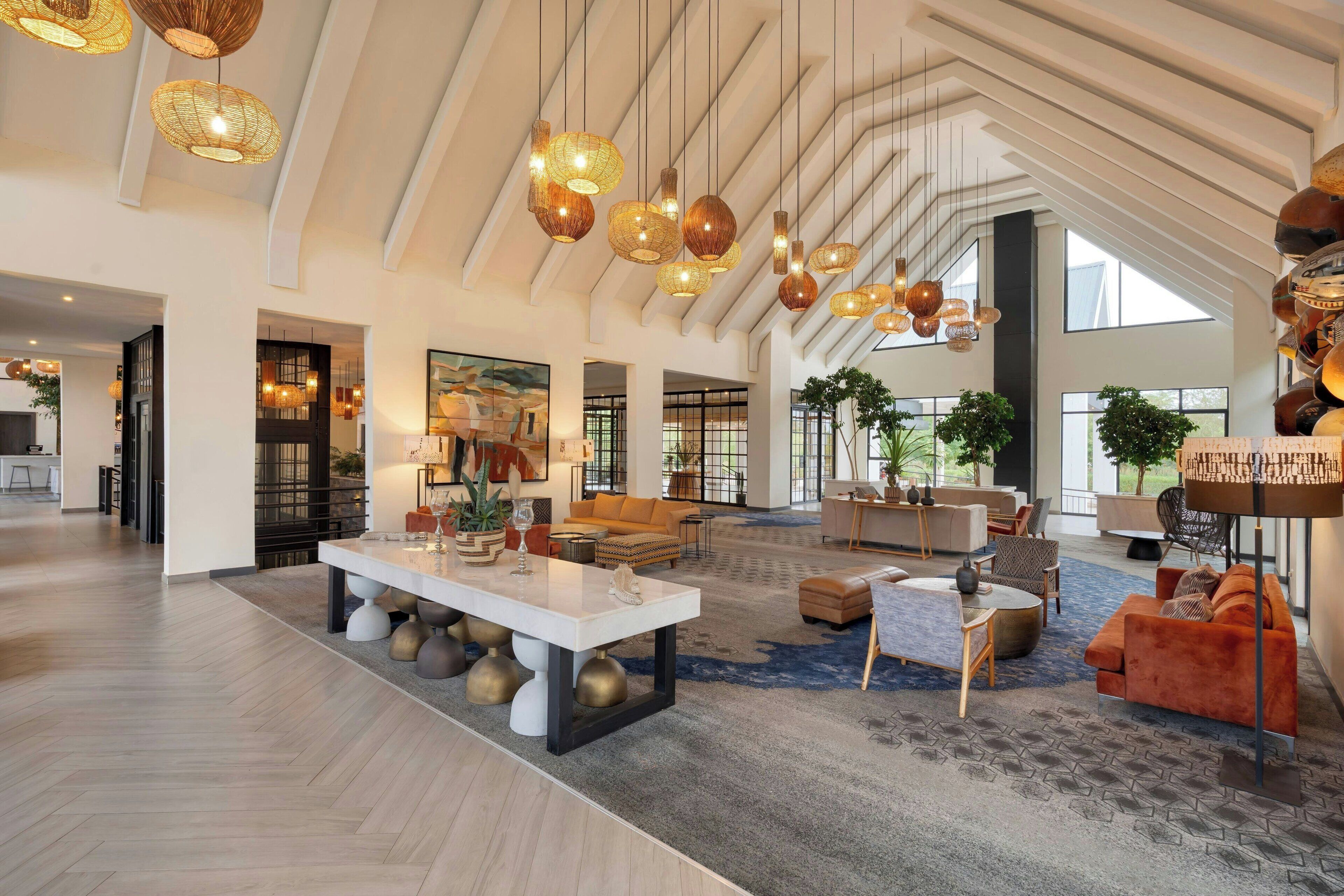Un hall d’entrée spacieux avec un plafond haut, des luminaires modernes, des coins salon confortables et de grandes fenêtres laissant entrer la lumière naturelle.