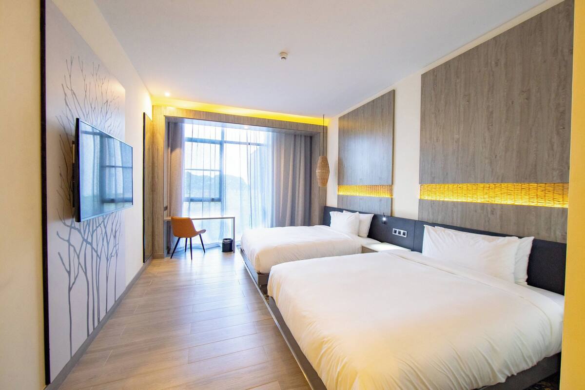 Ein Hotelzimmer mit zwei Betten, einem an der Wand befestigten Fernseher, einem Schreibtisch mit Stuhl und einem großen Fenster mit Vorhängen.