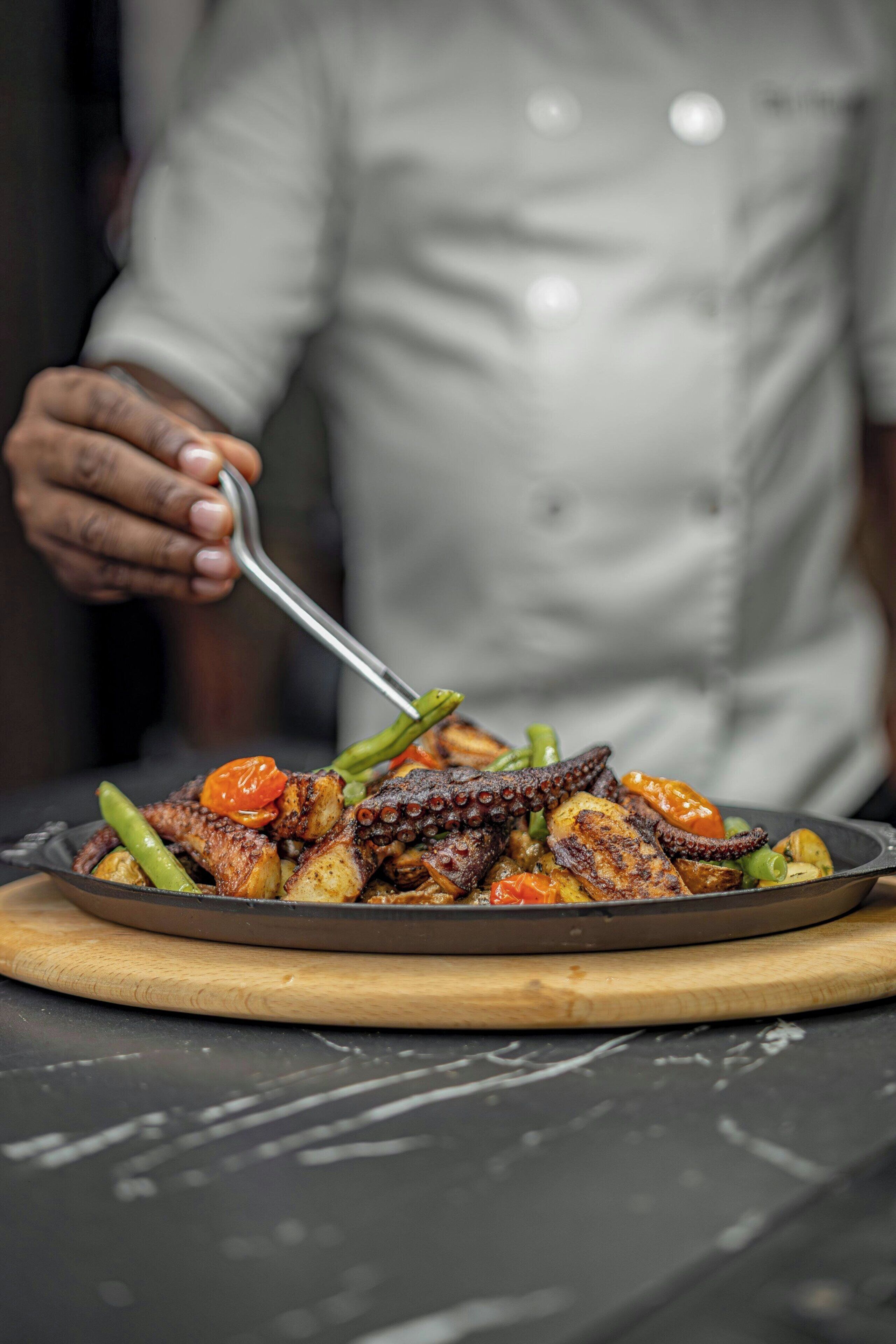 A chef serving a dish with octopus, potatoes, and vegetables.