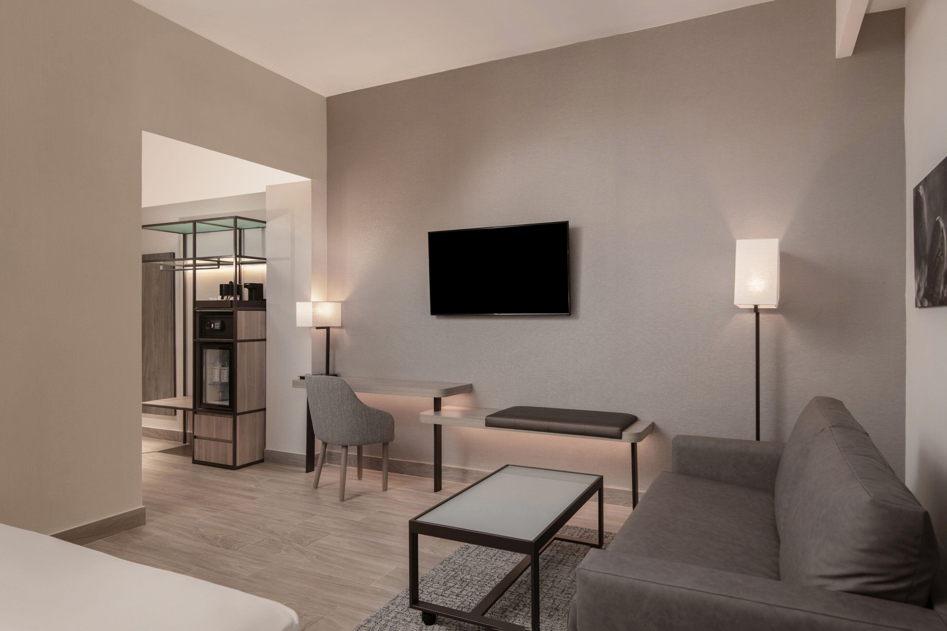 Una habitación de hotel moderna con sofá, una mesita, televisor montado en la pared y una zona de estar con una silla y un banco.