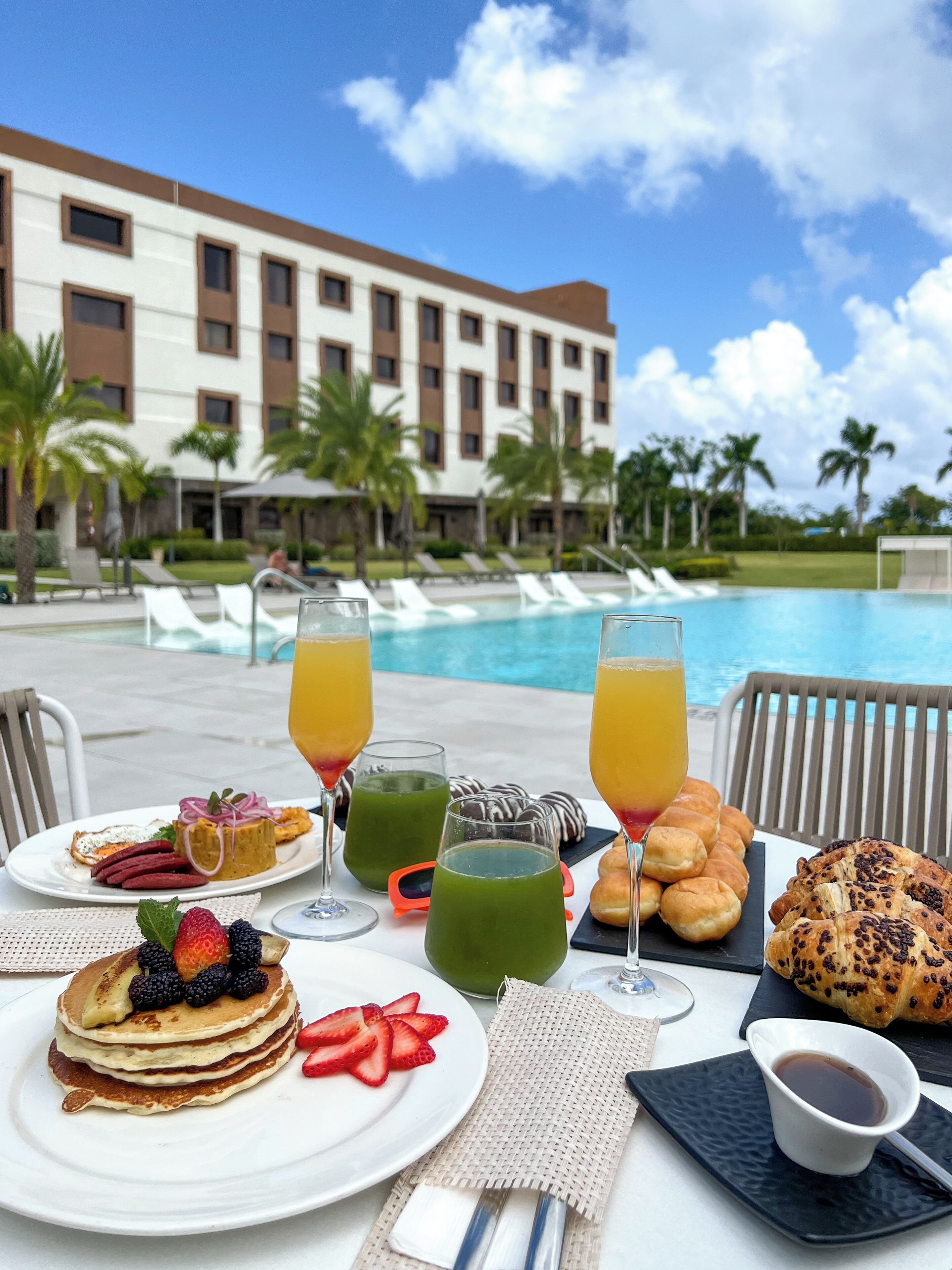 Une table de petit-déjeuner avec des crêpes, des fruits et des boissons, placée au bord d’une piscine, avec le bâtiment de l’hôtel en arrière-plan.
