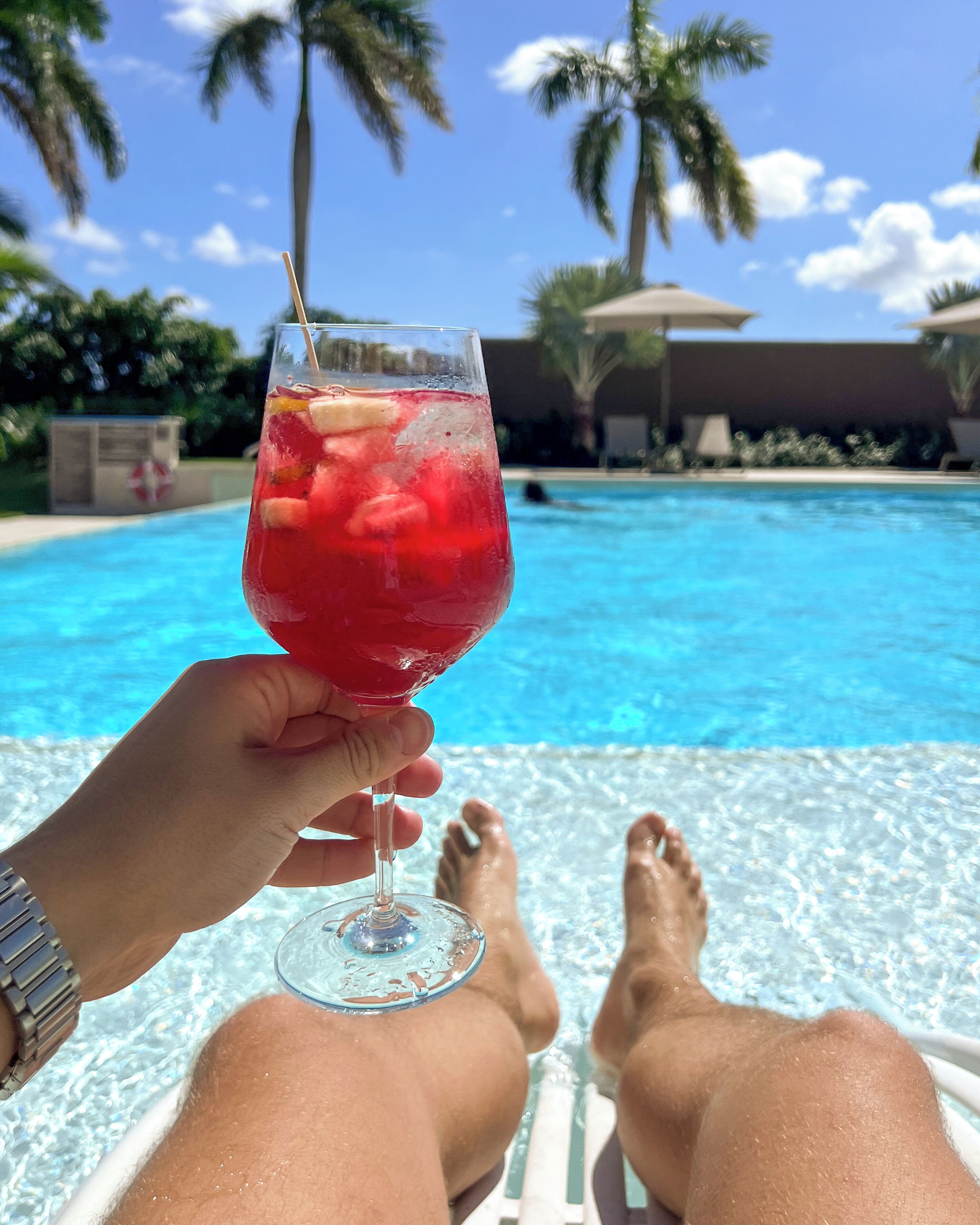 A person holding a glass of red drink by a poolside with clear blue water and palm trees in the background.