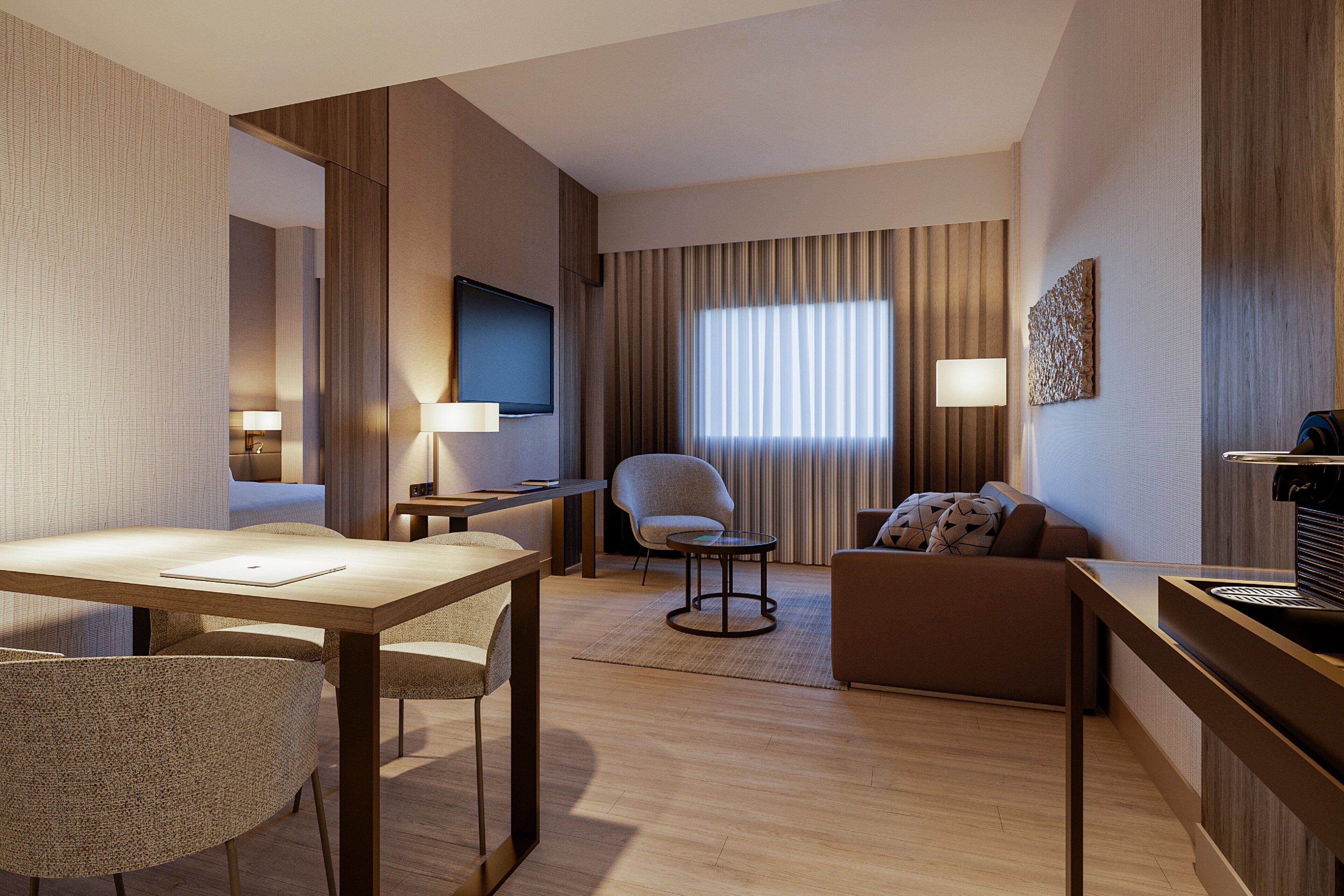 Une chambre d’hôtel avec un plancher en bois, une table à manger avec des chaises, un canapé, un téléviseur fixé au mur et une fenêtre avec des rideaux.