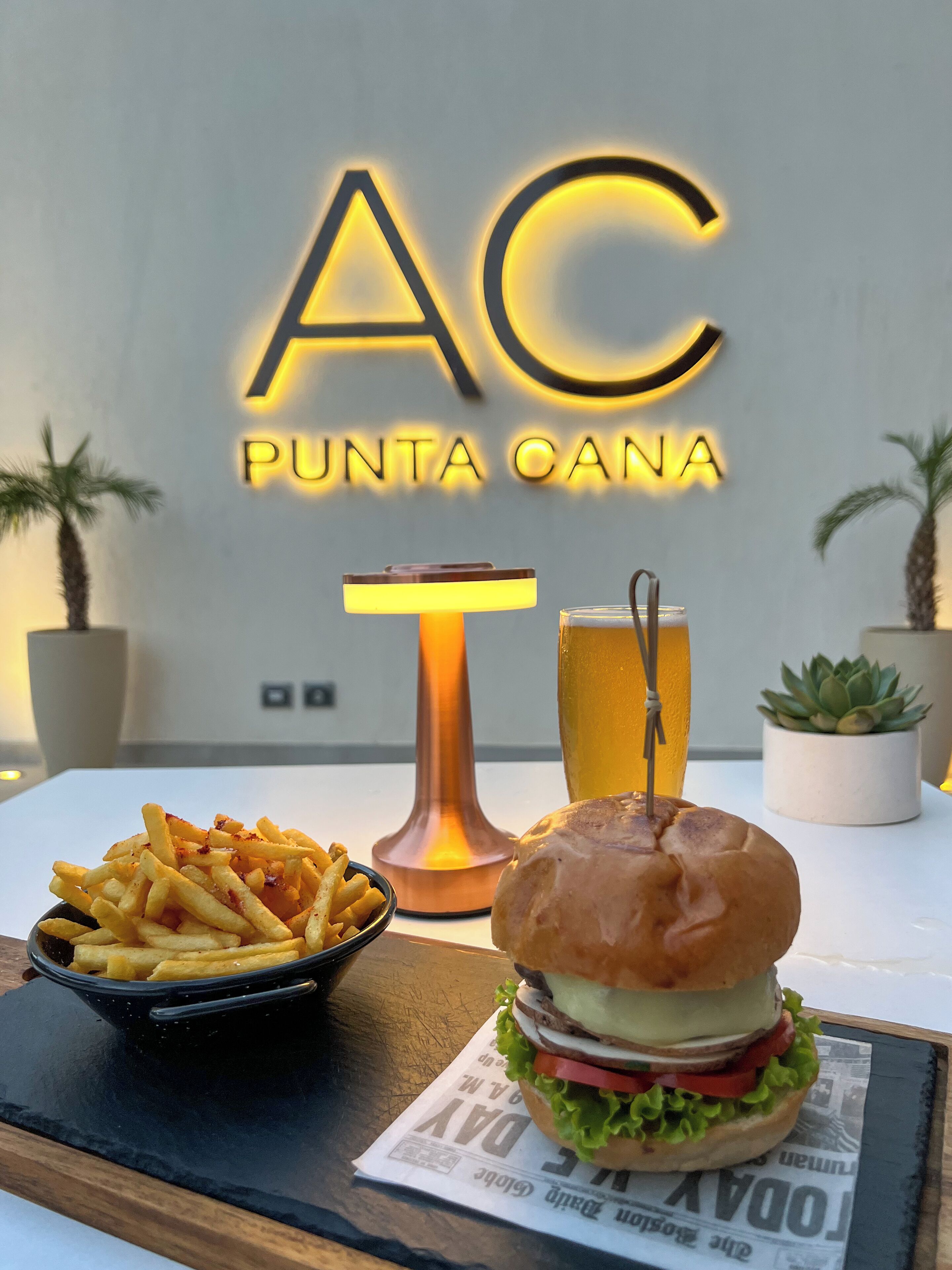 Una mesa con una hamburguesa, papas fritas y una cerveza, con un letrero de neón que dice "AC PUNTA CANA" al fondo.