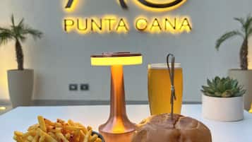 Una mesa con una hamburguesa, papas fritas y una cerveza, con un letrero de neón que dice "AC PUNTA CANA" al fondo.