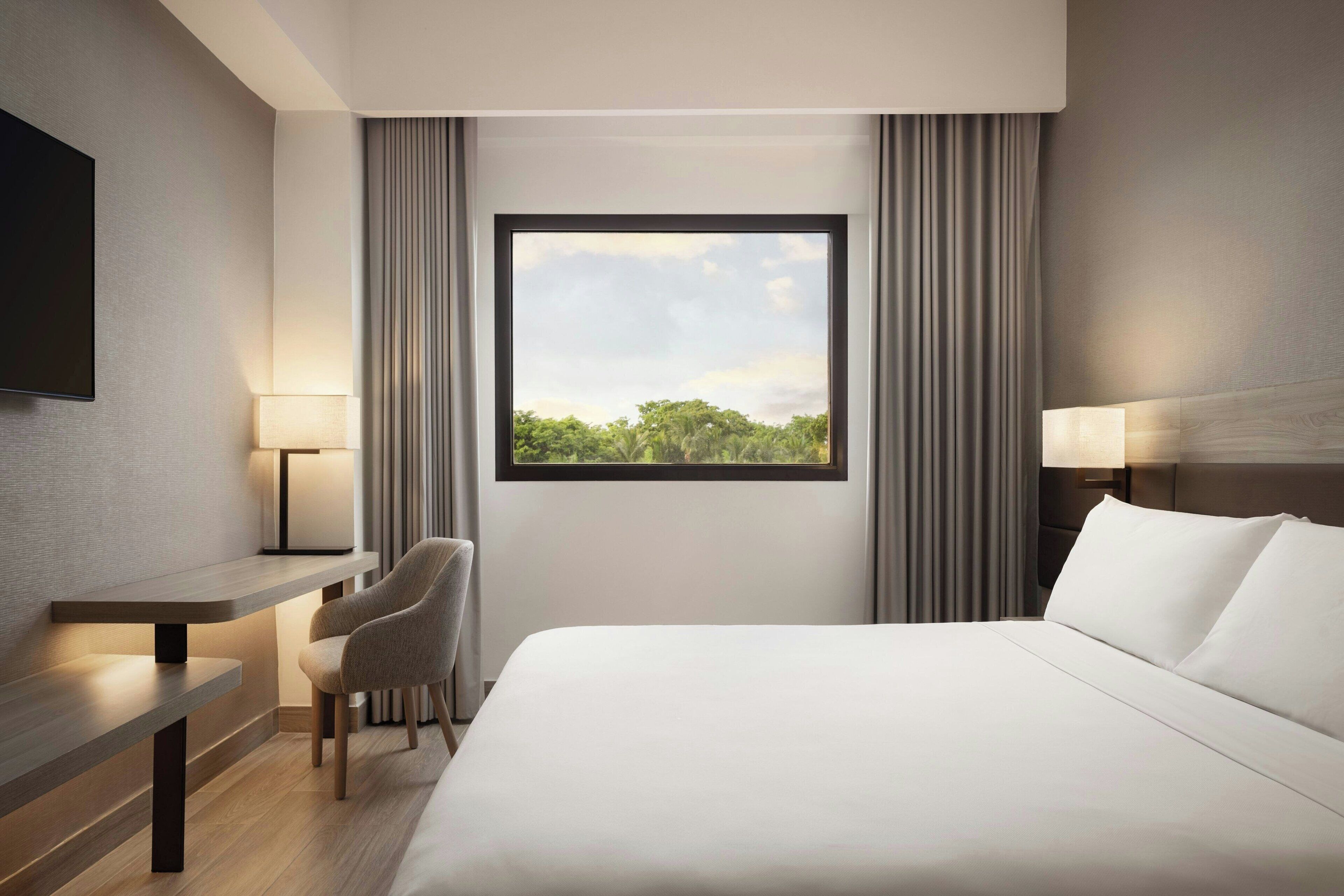 A hotel room with a bed, a desk, a chair, a TV, and a window with a view of trees.