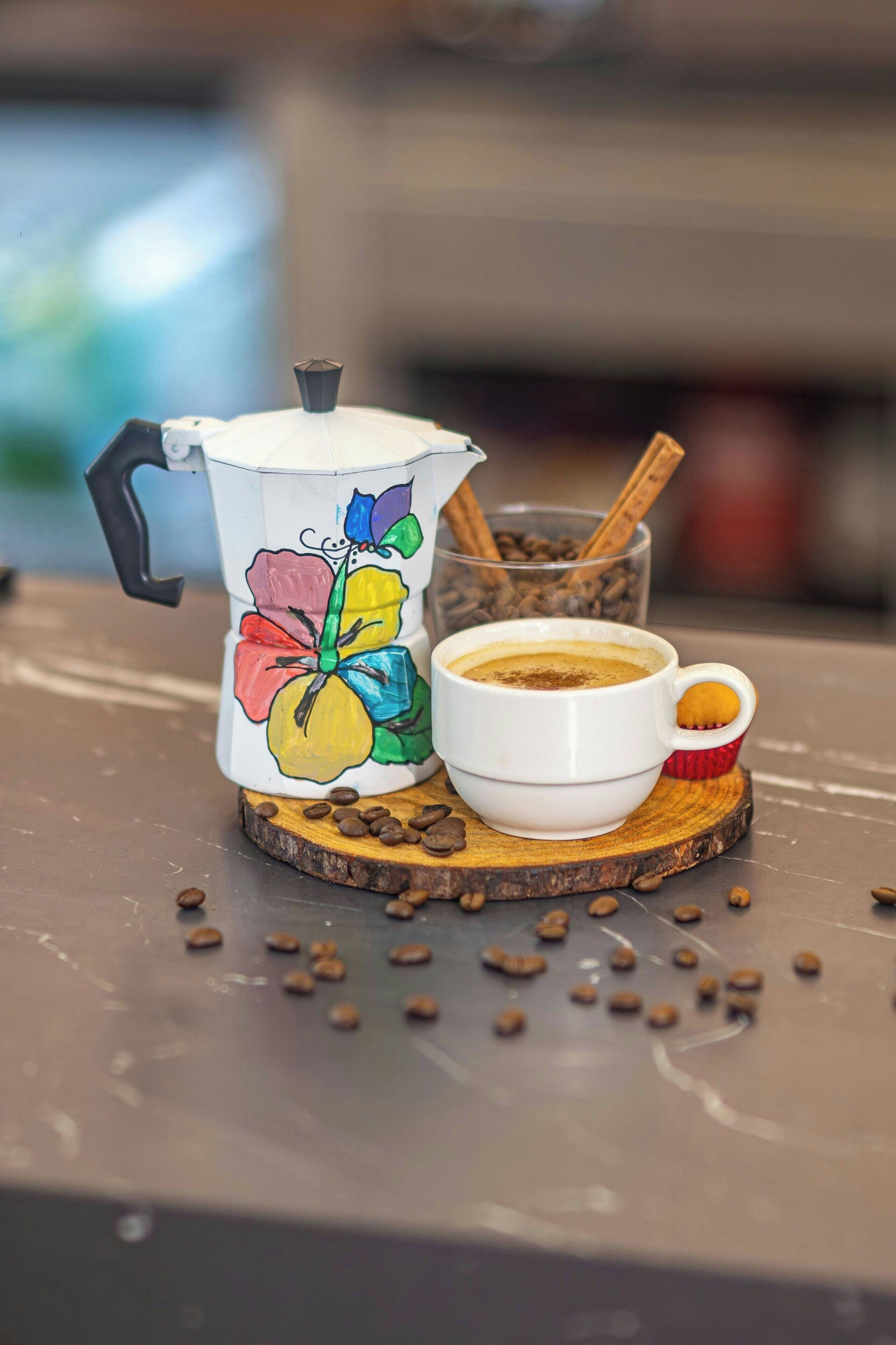 A coffee maker with a colorful floral design, a cup of coffee, and a cinnamon stick on a wooden coaster.